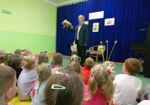 Sala gimnastyczna w przedszkolu. Dzieci siedą na widowni. Z uwagą patrzą na Eugeniusza Dolata, recytujacego jeden ze swoich wierszy i trzymającego pluszaka hipopotama.
