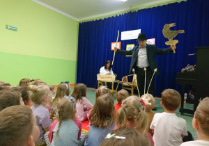 Sala gimnastyczna w przedszkolu. Dzieci siedą na widowni. Z uwagą patrzą na Eugeniusza Dolata w kapeluszu, recytujacego jeden ze swoich wierszy, trzymajacego w ręku kij i sylwetę ptaka .