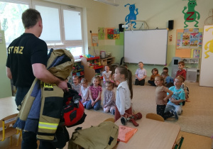 Sala przedszkolna. Strażak stoi przed siedzącymi na dywanie dziećmi, prezentuje sprzęt i wyposażenie strażaka.
