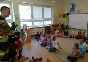 Sala przedszkolna. Strażak stoi przed siedzącymi na dywanie dziećmi, prezentuje sprzęt i wyposażenie strażaka.