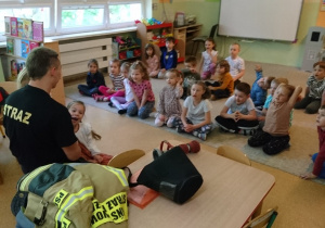 Sala przedszkolna. Strażak klęczy przed siedzącymi na dywanie dziećmi, opowiada o pracy strażaka.
