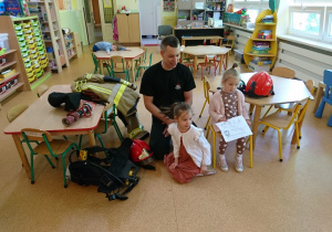 Sala przedszkolna. Strażak klęczy przed siedzącymi na dywanie dziećmi, opowiada o pracy strażaka.