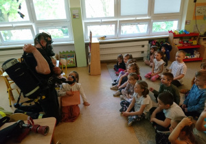 Sala przedszkolna. Strażak klęczy przed siedzącymi na dywanie dziećmi, prezentuje sprzęt do oddychania np. w pomieszczeniach zadymionych.
