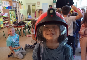 Sala przedszkolna. Chłopiec w hełmie strażackim na głowie, uśmiecha się.