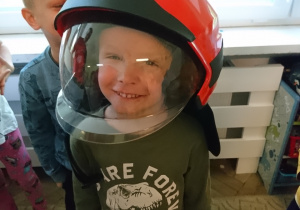 Sala przedszkolna. Chłopiec w hełmie strażackim na głowie, uśmiecha się.