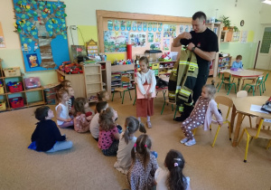 Sala przedszkolna. Strażak stoi przed siedzącymi na dywanie dziećmi i prezentuje sprzęt i wyposażenie strażaka.