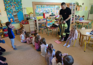 Sala przedszkolna. Strażak stoi przed siedzącymi na dywanie dziećmi. Pomaga dziewczynce założyć strażacki strój.
