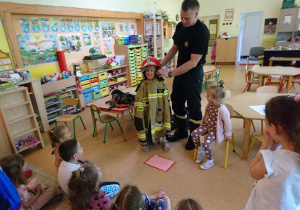 Sala przedszkolna. Strażak stoi przed siedzącymi na dywanie dziećmi. Strażak zakłada dziewczynce w stroju strażaka hełm strażacki.