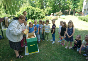 Ogród przedszkolny. Pani prowadząca warsztaty o pszczołach, stoi na przeciw dzieci. Prezentuje elementy ubioru pszczelarza. Obok niej stoi ul. Pani prezentuje elementy, z których zbudowany jest ul. Dzieci przyglądają się z uwagą.