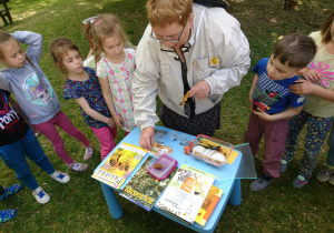 Ogród przedszkolny. Pani prowadząca warsztaty o prezentuje ciekawe publikacje, niezbędne w pracy pszczelarza. Dzieci przyglądają się z uwagą.