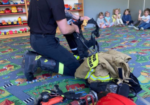 Strażak pokazuje dzieciom na dywanie osprzęt i ubranie strażaka