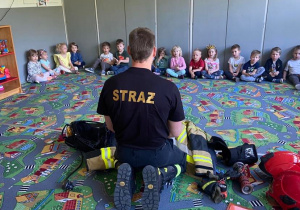 Dzieci siedzące na dywanie. Przed nimi strażak opowiadający o swojej pracy