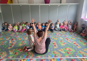 Dzieci na dywanie a przed nimi Mama dziewczynki z grupy. Wszyscy podnoszą ręce do góry