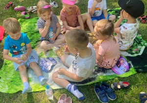 Dzieci siedzą na kocu w ogrodzie przedszkolnym. Spożywają posiłki.