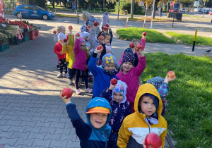 Dzieci ustawieni w parach na chodniku podnoszą do góry zakupione jabłka