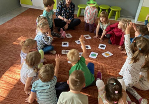 Dzieci na dywanie słuchają zwrotów w języku niemieckim pokazywanych na kartach obrazkowych przez nauczycielkę