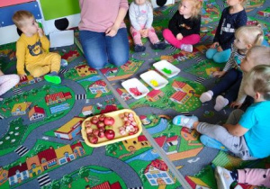 Dzieci siedzą w kole na dywanie. Przed nimi stoi taca z jabłkami i tacki z farbami. Nauczycielka pokazuje dzieciom szablon jabłoni