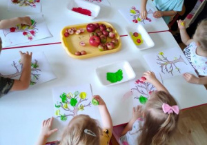 Dzieci stemplują połówkami jabłek na kartkach z szablonem jabłoni
