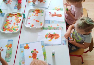 Dzieci przyklejają kolorowe jabłuszka na kartonie z odbitym z dłoni drzewkiem