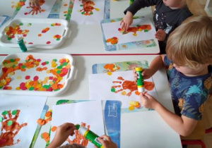 Dzieci przyklejając małe jabłuszka na kartki z drzewami utworzonymi z dłoni dzieci