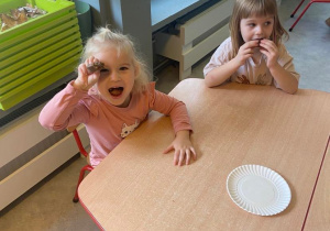 Dziewczynki przy stole jedzące czekoladowe babeczki, jedna z nich przykłada babeczkę do oka