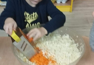 Sala przedszkolna. Chłopiec siedzi przy stoliku i trze marchewkę.