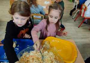 Sala przedszkolna. Dwie dziewczynki siedzą przy stoliku i wkładają kapustę do słoików