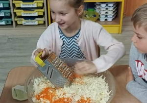 Sala przedszkolna. Dziewczynka siedzi przy stoliku i trze marchewkę