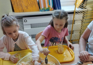 Sala przedszkolna. Dwie dziewczynki siedzą przy stoliku i zgodnie z instrukcją nauczycielki wypełniają słoiki poszatkowaną kapustą.