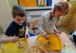 Sala przedszkolna. Dziewczynka i chłopiec siedzą przy stoliku i wkładają kapustę do słoików