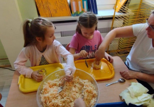 Sala przedszkolna. Dwie dziewczynki siedzą przy stoliku i zgodnie z instrukcją nauczycielki wypełniają słoiki poszatkowaną kapustą.