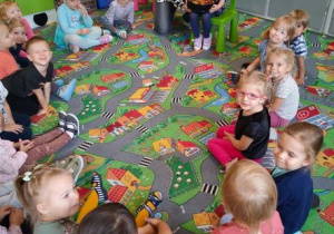 Dzieci siedzą w kole na dywanie. Nauczycielka wraz z jubilatką prezentują urodzinowy tort owocowy