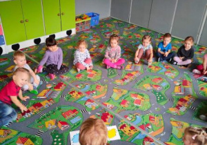 Dzieci siedzą w kole na dywanie wraz z nauczycielką. Na dywanie leżą flashcards przedstawiające kolory