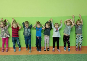 Dzieci stoją w szeregu plecami do ściany. Trzymając ramiona w górze stają na palcach