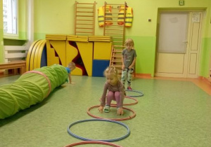 Dzieci wskakują do kolejno ułożonych w rzędzie obręczy. W tle chłopiec pokonuje tunel gimnastyczny