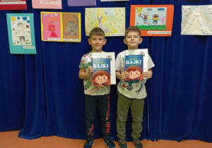 Chłopcy pozują do zdjęcia na tle wystawy prac plastycznych trzymając w rękach nagrody książkowe