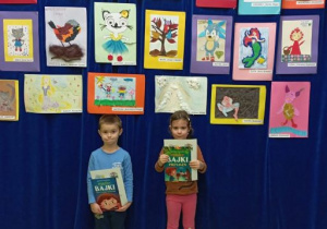 Chłopiec i dziewczynka pozują do zdjęcia na tle wystawy prac plastycznych trzymając w rękach nagrody książkowe