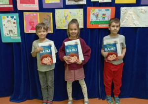 Dzieci pozują do zdjęcia na tle wystawy prac plastycznych trzymając w rękach nagrody książkowe