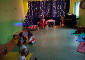 Mikołaj siedzi na fotelu obok choinki. Wskazuje ręką na prezenty leżące pod choinką. Przed Mikołajem siedzą dzieci