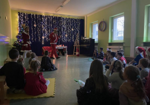 dzieci siedzą w sali wśród świątecznej scenografii i słuchają Mikołaja