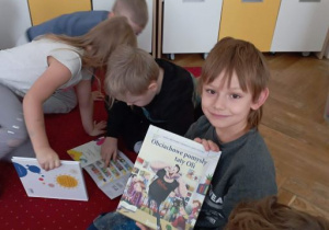 Chłopiec prezentuje okładkę jednej z książek. Obok chłopca dzieci oglądają książki
