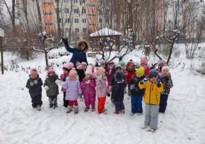 Dzieci z nauczycielką pozują do zdjęcia na tle zasypanego śniegiem ogrodu przedszkolnego