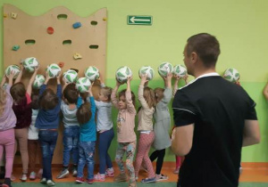 Dzieci trzymają piłki w górze. Trener obserwuje dzieci.