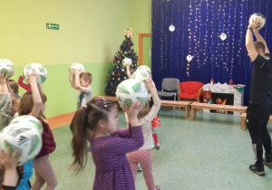 Dzieci chodzą po sali trzymając piłki nad głowami.