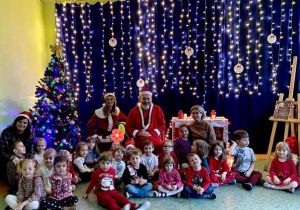 Dzieci siedzą na sali gimnastycznej, towarzyszą im Panie oraz Święty Mikołaj i Pani Mikołajkowa.