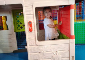 Dziewczynka uśmiecha się stojąc w okienku zabawkowego domku