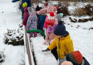 Nauczycielka pokazuje dzieciom pokryte śniegiem kwiaty. Kilkoro dzieci bawi się śniegiem pokrywającym ławkę