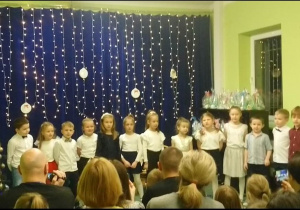 Sala gimnastyczna. Dzieci w odświętnych strojach stoją na tle dekoracji świątecznej. Z prawej strony znajduje się choinka, z lewej strony, widać upominki przygotowane przez dzieci. Przed dziećmi na widowni siedzą rodzice.