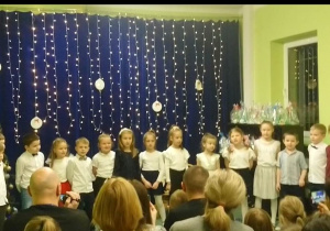 Sala gimnastyczna. Dzieci w odświętnych strojach stoją na tle dekoracji świątecznej. Z prawej strony znajduje się choinka, z lewej strony, widać upominki przygotowane przez dzieci. Przed dziećmi na widowni siedzą rodzice.