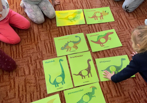 Dzieci na dywanie przed tablicami z przedstawiające dinozaury